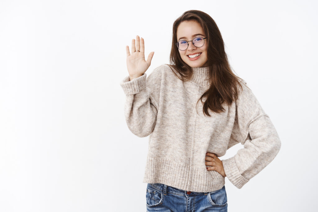 Silly pleasant and charismatic young woman in glasses and sweater flirting and laughing cute as raising hand in hi or hello gesture, waiting to greet person, smiling at camera over gray wall